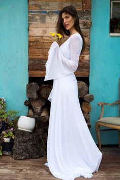 a woman in a long white dress holding a flower and standing next to a blue building