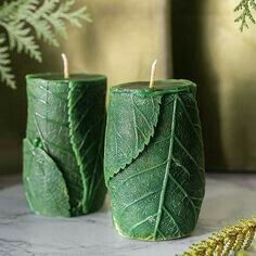 two green candles sitting next to each other on a marble table with greenery in the background