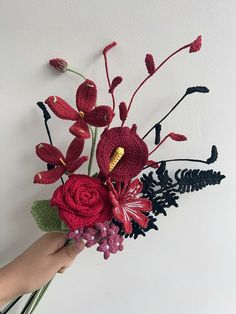 a hand holding a crocheted flower arrangement in front of a white wall with black and red flowers