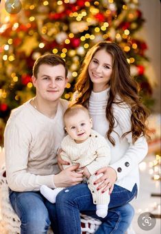 a young man and woman holding a baby in front of a christmas tree with lights