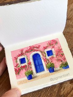 someone is holding up a card with a house painted on it and flowers in the window