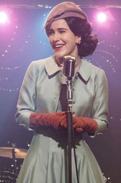 a woman in a blue dress and red gloves is holding a microphone while smiling at the camera