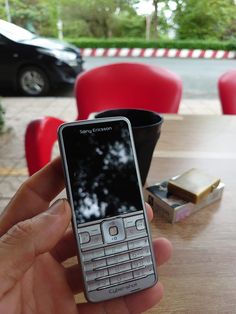 a hand holding a cell phone over a wooden table with red chairs in the background