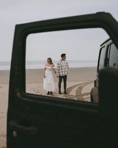 a man and woman standing on the beach in front of a car looking at each other