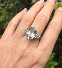 a woman's hand holding an oval shaped ring with two diamonds on it, in front of some bushes