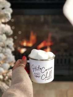 a woman holding a mug with marshmallows in it and a fireplace behind her