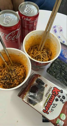 two bowls of noodles with chopsticks in them on a table next to cans of soda