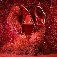 a heart - shaped object is surrounded by red plants and bushes in front of a wall