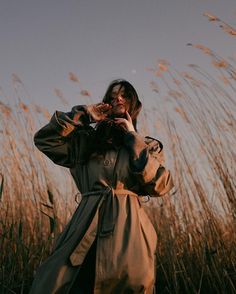 a woman standing in tall grass talking on a cell phone while wearing a trench coat