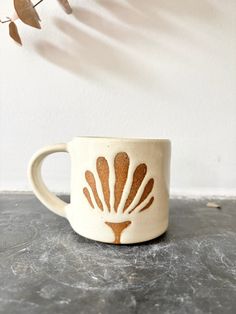 a white and brown coffee cup sitting on top of a gray table next to a plant