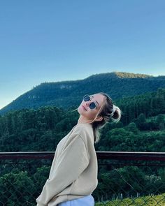 a woman wearing sunglasses is sitting on a fence and looking up at the sky with mountains in the background