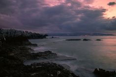 the sun is setting over the ocean with rocks in the foreground and clouds in the background