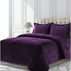 a bed with purple comforter and pillows in a white room next to a lamp