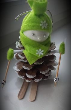 a green stuffed animal sitting on top of a pine cone and holding two wooden sticks