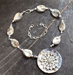 "This stunning necklace incorporates a beautiful, oversize clear rhinestone brooch, set into an even larger Paua shell button. The Paua shell backing for the brooch is 2 7/8\" in diameter. The necklace measures 18 1/2\" from clasp end to clasp end. I have also included a very pretty 5\" extension chain that is removable. The beads are of matching shell and are freeform. So beautiful. Each beaded segment is wire-wound to the next." Unique Large Pendant Jewelry For Wedding, Unique Large Pendant Wedding Jewelry, Unique Mother Of Pearl Jewelry For Wedding, Unique Silver Bridal Necklace, Unique Silver Bridal Necklace For Wedding, Wedding Pendant Necklace With Bling, Unique Jeweled Necklaces For Wedding, Unique Rhinestone Wedding Jewelry, Vintage Mother Of Pearl Jewelry For Wedding