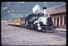 an old fashioned steam train is on the tracks near a building with mountains in the background