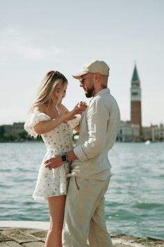 a man and woman standing next to each other near the water with a clock tower in the background