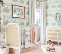 a baby sitting on the floor next to a crib in a room with floral wallpaper