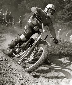 a man riding on the back of a motorcycle down a dirt road while people watch