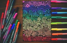 some markers and pens are next to a notebook with an intricate design on it, surrounded by other colored pencils