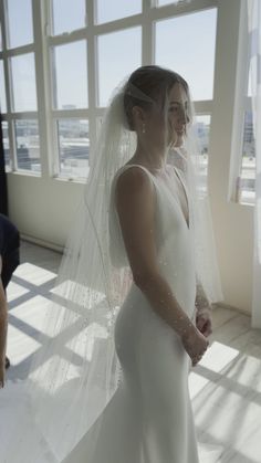 a woman in a white wedding dress is standing by a window with her veil pulled back