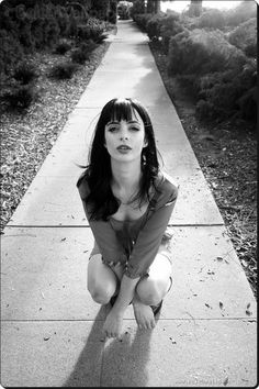 black and white photograph of a woman sitting on the sidewalk with her hands in her pockets