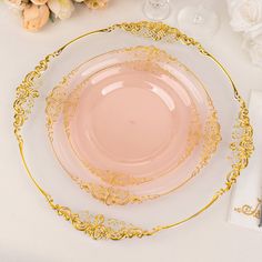 a pink plate with gold trim sits on a white tablecloth next to some wine glasses and flowers