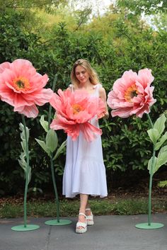 a woman is standing in front of three large pink flowers with green stems and leaves