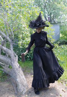 a woman in a black dress and hat standing next to a tree