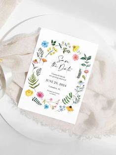 a wedding card with watercolor flowers and leaves on it next to a white plate