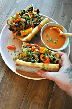 a person holding a plate with two sandwiches on it and dipping sauce in the bowl