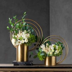 two gold vases with flowers and greenery in them on top of a table