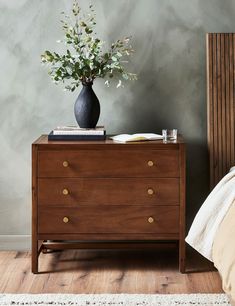 a vase with flowers on top of a wooden dresser next to a bed in a bedroom