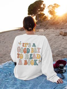 a woman sitting on top of a blue blanket next to a tree and ocean with the words it's a good day to read a book