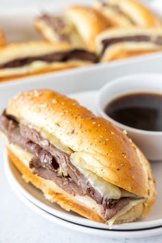 a roast beef sandwich on a plate with dipping sauces and bread rolls in the background