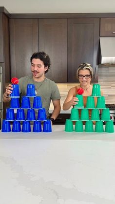 two people standing behind a kitchen counter with blue and green cups in front of them