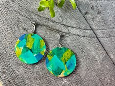 two circular earrings with green and blue designs on them sitting on a wooden surface next to a potted plant