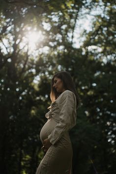 a pregnant woman standing in front of trees with the sun shining through her hair and wearing a coat