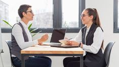 two people sitting at a table with laptops in front of them, one is talking to the other