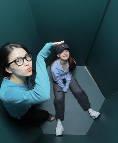 two young women sitting on the floor in front of a green wall, one pointing at something