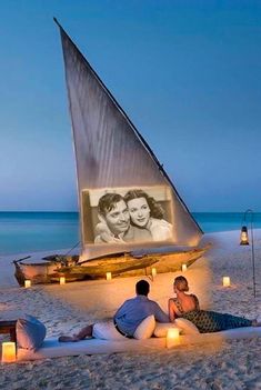 two people sitting on the beach with candles in front of them and a sailboat