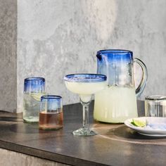 three glasses and pitcher sitting on top of a counter next to a plate with lime slices