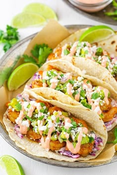 three fish tacos on a plate with lime wedges and cilantro sauce