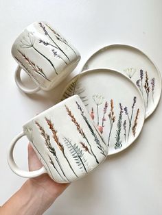 a hand holding three coffee cups and saucers with plants painted on the sides, in front of a white background