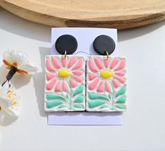 a pair of pink and green flower earrings on top of a white card next to flowers