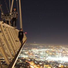 two people standing on the edge of a tall building at night with city lights in the background