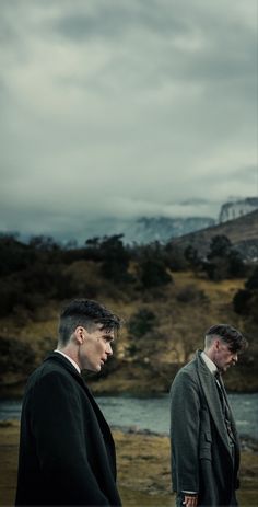 two men standing next to each other in front of a lake and mountains on a cloudy day