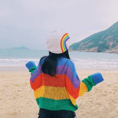 a woman standing on top of a sandy beach next to the ocean wearing a rainbow sweater