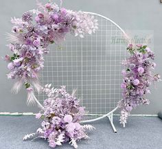 purple flowers and greenery are arranged around a white circular frame on the floor in front of a gray wall