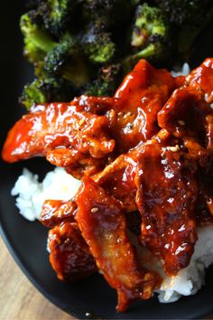 a black plate topped with meat covered in sauce and rice next to broccoli